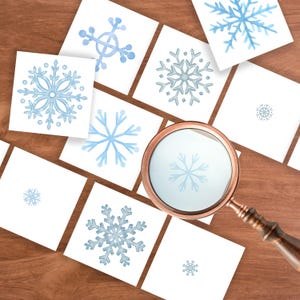 May include: A collection of white square cards with watercolor snowflake designs in various shades of blue and gray. A magnifying glass with a copper-colored frame and wooden handle is positioned over the cards, magnifying a snowflake image.