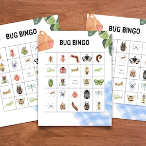 May include: Three Bug Bingo game cards with watercolour illustrations of various insects. The cards feature a grid layout with images of bugs and the text "BUG BINGO" at the top. A picnic basket and greenery are also depicted.