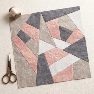 Könnte beinhalten: Ein quadratischer Stoff-Quiltblock mit einem geometrischen Muster aus Dreiecken und Trapezen in Altrosa, Anthrazit, Weiß und Beige mit Punkt- und Blumenmustern. Eine Garnrolle und eine Metallschere liegen neben dem Stoffstück auf dem Untergrund.