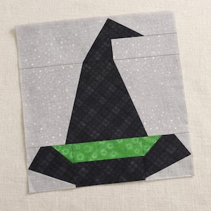 May include: A square fabric quilt block featuring a pieced witch hat design. The hat is constructed from black fabric with a subtle grid pattern, accented by a bright green band. The background is a light gray fabric with a white speckled pattern.