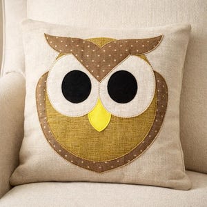 May include: A square scatter cushion with a beige linen-textured cover featuring a large owl appliqué. The owl has brown polka-dot ears, large white circular eyes with black pupils, a mustard yellow textured body, and a bright yellow triangular beak.