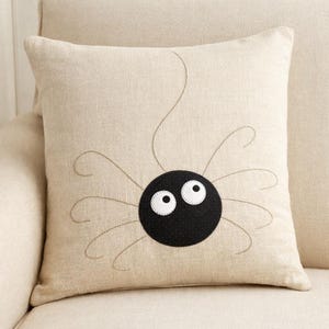 May include: A square beige throw pillow featuring a centered black circular spider body with two large white eyes. The spider has eight thin, curved legs embroidered in a dark thread, extending outward from the central black circular shape.