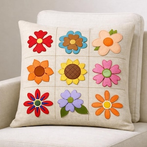 May include: A square beige throw pillow featuring a grid of nine distinct felt flower appliqués in various colors including red, blue, orange, yellow, pink, purple, and peach. Each flower is centered within its own stitched square section.