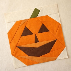 Puede incluir: Un bloque de edredón cuadrado de tela con un diseño de calabaza. La calabaza está construida con varios tonos de tela naranja con ojos triangulares de color marrón oscuro, una nariz marrón y una boca ancha de color marrón.