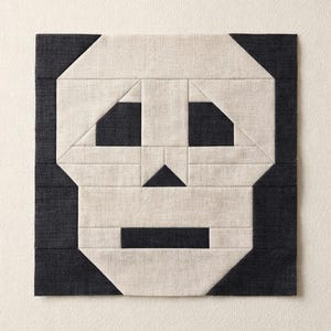 May include: A square quilt block featuring a stylised skull design constructed from fabric pieces. The skull is composed of light beige fabric for the face and dark charcoal grey fabric for the eyes, nose, mouth, and corners of the square block.
