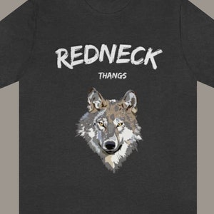 May include: A dark gray t-shirt with the text "REDNECK THANGS" in white paint-like lettering. A wolf's head is centered below the text. The text "REDNECK THANGS" is also visible in red lettering at the bottom of the image.