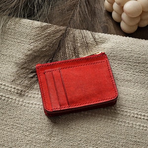 May include: A red suede cardholder with three card slots and a zippered compartment. The wallet is rectangular and has a gold-colored zipper. It rests on a textured, neutral-colored surface.