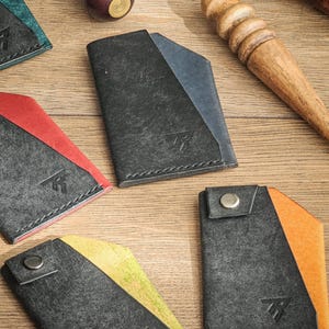 Könnte beinhalten: Eine Sammlung handgefertigter Ledergeldbörsen in verschiedenen Farben, darunter Schwarz, Blau, Rot und Orange. Jede Geldbörse hat ein einzigartiges Design mit einem kontrastierenden Farbakzent und einem Metall-Druckknopfverschluss. Die Geldbörsen werden auf einer Holzoberfläche präsentiert.