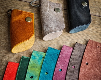 May include: A set of nine leather keyrings in various colours, including brown, tan, black, green, blue, red, purple, and grey. The keyrings are made of leather and have a metal ring for attaching keys. The keyrings are arranged in a row on a wooden surface.