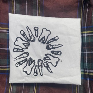 May include: A white canvas pouch with a black illustration of teeth in a circular design. The pouch is rectangular and rests on a plaid fabric background with brown, green, and blue hues.