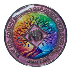 Pode incluir: Um medalhão de recuperação circular de metal com um design de árvore em cores de arco-íris. Um losango central contém as letras NA. A borda externa é rosa metálico com o texto THAT NO ADDICT SEEKING RECOVERY NEED EVER DIE e 1991 NAWS.
