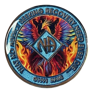 Op de afbeelding: Een blauwe en zilveren munt met een feniks die uit vlammen opstijgt. De munt heeft de tekst "THAT ADDICT SEEKING RECOVERY NEED EVER DIE" rond de rand en "NA" in een ruitvorm in het midden. De munt heeft ook de tekst "©1991 NAWS" onderaan.