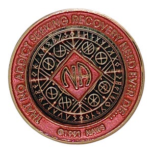 May include: A circular red and gold metal medallion featuring the letters NA in a central diamond. The border text reads THAT NO ADDICT SEEKING RECOVERY NEED EVER DIE and 1991 NAWS. The design includes geometric patterns and runic-style symbols.