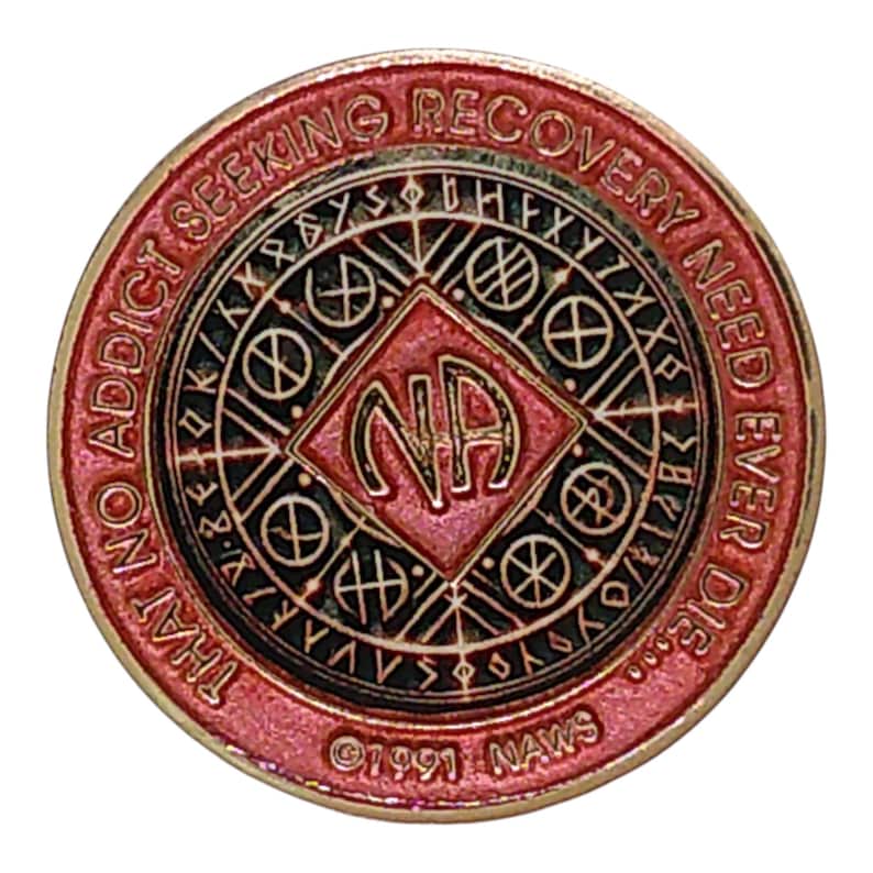 May include: A circular red and gold metal medallion featuring the letters NA in a central diamond. The border text reads THAT NO ADDICT SEEKING RECOVERY NEED EVER DIE and 1991 NAWS. The design includes geometric patterns and runic-style symbols.