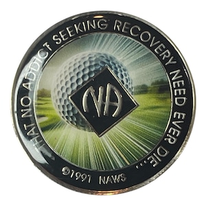 Könnte beinhalten: Eine silberne Münze mit schwarzem Hintergrund und einem weißen Golfball mit einem schwarzen NA-Logo in der Mitte. Der Text am Rand lautet "That no addict seeking recovery need ever die... 1991 NAWS".