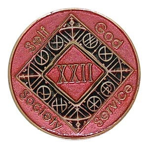 May include: A round pink and gold metal medallion featuring a central diamond shape with the Roman numeral XXII inside. The words Self, God, Society, and Service are embossed around the perimeter, with black accents filling the geometric patterns.