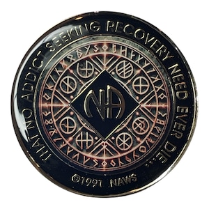 Può includere: Una moneta di metallo dorata e nera con le parole "I am no addict seeking recovery need ever did" e "NAWS" attorno al bordo. Il centro della moneta presenta un quadrato nero con le lettere "NA" in oro e un cerchio di simboli intorno.