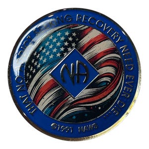 May include: A circular metal recovery medallion featuring a blue border with the text THAT NO ADDICT SEEKING RECOVERY NEED EVER DIE and 1991 NAWS. The center displays a stylized American flag design with a blue diamond containing the letters NA.