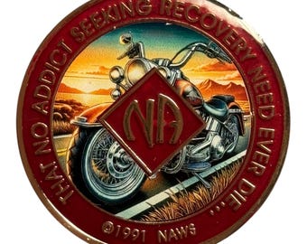 May include: A round metal medallion featuring a red border with the text THAT NO ADDICT SEEKING RECOVERY NEED EVER DIE. The center depicts a motorcycle on a road at sunset with a red diamond overlay containing the gold letters NA and 1991 NAWS.