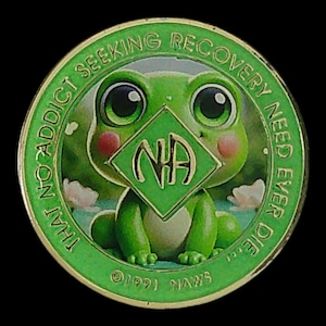 May include: A round green metal token featuring a cartoon frog with large eyes and pink cheeks. A central green diamond displays the letters NA. The border text reads THAT NO ADDICT SEEKING RECOVERY NEED EVER DIE. The bottom includes 1991 NAWS.