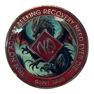 Puede incluir: Un medallón redondo con un borde rojo oscuro que presenta el texto THAT NO ADDICT SEEKING RECOVERY NEED EVER DIE. El centro muestra una ilustración de un dragón detrás de un rombo rojo con las letras NA. La parte inferior incluye 1991 NAWS.