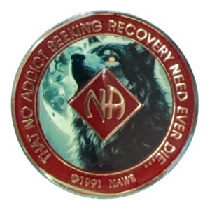 May include: A circular metal medallion with a red border featuring the text THAT NO ADDICT SEEKING RECOVERY NEED EVER DIE and 1991 NAWS. The center displays a wolf image behind a red diamond shape containing the gold letters NA.