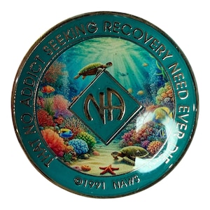 May include: A teal and silver coin with an underwater scene featuring turtles, coral, and fish. The coin has the letters "NA" inside a diamond and the text "THAT NO ADDICT SEEKING RECOVERY NEED EVER DIE". The bottom of the coin reads "©1991 NAWS".