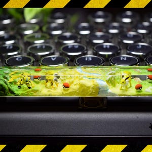 May include: A green, yellow and brown resin keycap with a Pokemon theme. The keycap features a landscape with small Pokemon figures and Pokeballs. The keycap is sitting on top of a black keyboard with silver keys.