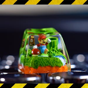 May include: A clear resin keycap featuring a miniature Mario character. The keycap depicts a green and brown landscape with a mushroom and a pipe.
