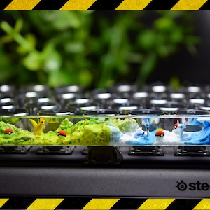 May include: A custom keyboard with a clear resin spacebar featuring miniature Pokémon figures and landscapes. The design includes a Pikachu, a blue wave, and red and white Poké Balls, set against a backdrop of green foliage.