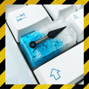 May include: A custom keycap with a black and silver kunai knife design set in a resin block with blue and white swirls, on a white keyboard. This keycap is a unique accessory for mechanical keyboards.