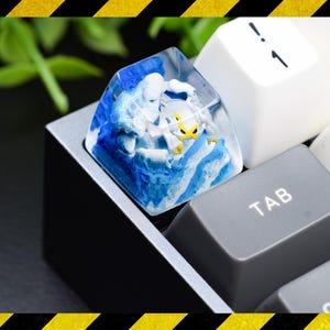 May include: A custom artisan keycap featuring a clear resin housing with a blue and white swirling wave pattern. Inside the keycap is a small, detailed white and yellow creature figure. The keycap is installed on a mechanical keyboard next to a grey TAB key.