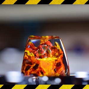Pode incluir: Um keycap de resina vermelho e laranja com uma pequena figura de dragão vermelho dentro. O dragão é cercado por resina laranja e amarela que se assemelha a chamas.