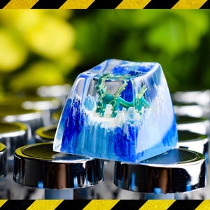 Peut inclure: Une touche de clavier artisanale personnalisée avec une scène de dragon miniature encapsulée dans de la résine transparente. La touche présente des motifs bleus et blancs en forme de vagues, avec une figurine de dragon vert à l'intérieur.