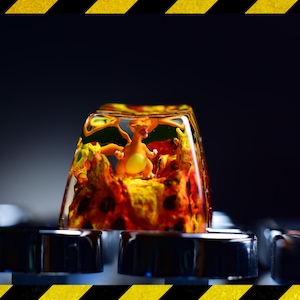 May include: A clear resin keycap with a small orange and yellow figure inside. The figure is surrounded by orange and yellow resin, creating a fiery effect. The keycap is sitting on a silver keyboard.