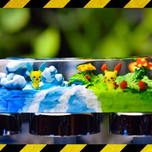 May include: A clear resin keycap with a miniature scene inside. The scene features a blue and white water-like landscape on the left, a green grassy landscape on the right, and two yellow Pokemon characters in the middle.