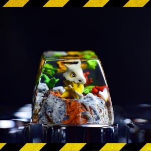 May include: A custom keycap with a Cubone figure set in a miniature landscape. The clear keycap showcases the figure and landscape within. The landscape includes rocks, plants, and colourful accents. The keycap is designed for use on a keyboard.