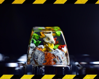Può includere: Un copritasto personalizzato con un personaggio di Cubone in un paesaggio in miniatura. Il copritasto trasparente mostra la figura e il paesaggio all'interno. Il paesaggio include rocce, piante e accenti colorati. Il copritasto è progettato per una tastiera.
