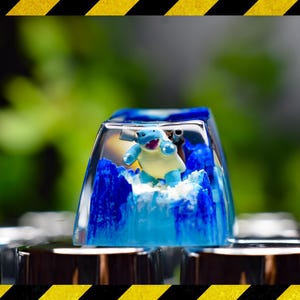 May include: A blue and white resin keycap with a small blue Pokemon character inside. The keycap is sitting on a silver surface.