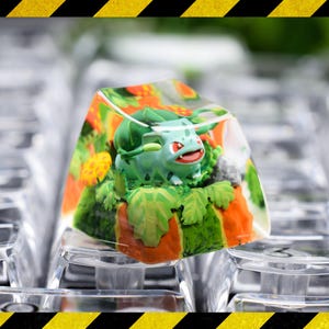 May include: A clear resin mechanical keyboard keycap featuring a miniature green Bulbasaur figure surrounded by sculpted orange and green foliage, including small mushrooms, encased within the transparent material of the keycap structure.