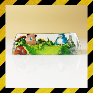 May include: A clear resin art piece featuring two cartoon characters in a miniature landscape. One character is pink with black ears, and the other is blue and white. The scene includes green foliage, mushrooms, and a rock formation.