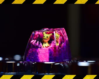 Puede incluir: Un tapón de tecla de resina transparente con un diseño de remolino rosa y morado. Dos figuras de dibujos animados amarillas son visibles dentro de la resina.