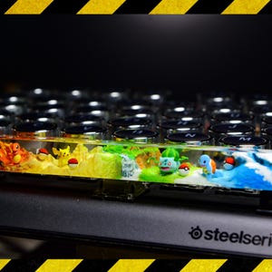 Könnte beinhalten: Eine benutzerdefinierte Tastaturkappe mit einem klaren Harzdesign, das verschiedene Pokémon-Charaktere zeigt, darunter Glumanda, Bisasam, Schiggy und Pikachu. Die Tastaturkappe ist vor einem schwarzen Hintergrund mit einem SteelSeries-Logo platziert.