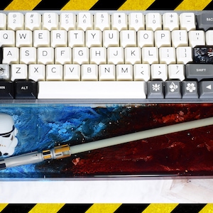 Puede incluir: Un teclado mecánico blanco y gris con un conjunto de teclas personalizadas con temática de Star Wars. El teclado está apoyado en una alfombrilla de ratón de resina azul y roja con un sable de luz blanco y una figura de Stormtrooper blanca.