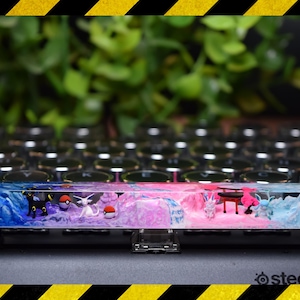 Puede incluir: Un tapón de tecla de teclado personalizado con un diseño de resina azul y rosa con personajes de Pokémon y un templo japonés. El tapón de tecla está hecho de resina transparente con un patrón azul y rosa en espiral. Los personajes de Pokémon son visibles en la resina, incluyendo un Eevee blanco y negro, un Sylveon rosa y blanco y un Glaceon azul y blanco. El templo japonés es una pequeña estructura roja con un techo blanco.