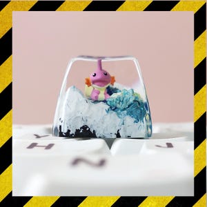 May include: A custom keycap with a pink and orange cartoon character encased in clear resin. The keycap has a white and blue base, creating a miniature underwater environment. The keycap is on a white keyboard.