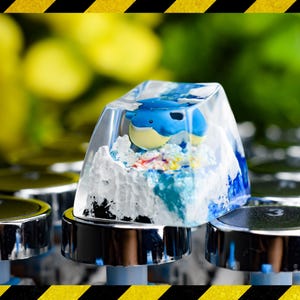 May include: A custom keycap with a blue and yellow cartoon character inside a clear resin block, resembling ice. The keycap is mounted on a silver and black mechanical keyboard switch. The background is blurred green and yellow.