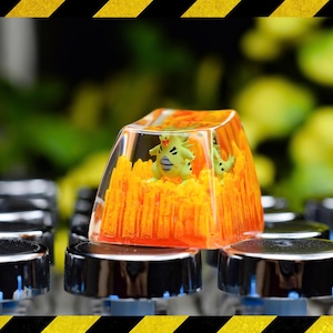May include: A custom keycap with a green and yellow creature inside a clear resin block with orange detailing. The keycap sits on a black keyboard with silver key bases. The background is a blurred green.