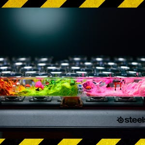 May include: A clear, rectangular resin keycap with a colourful, multi-layered design. The keycap features a landscape scene with red, green, and pink layers, and small figurines. The keycap is resting on a black keyboard with the SteelSeries logo visible.