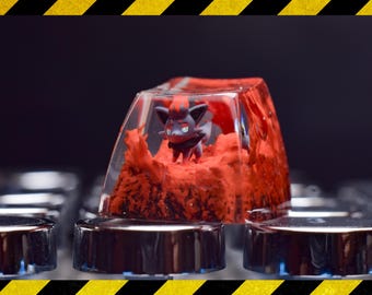 Può includere: Un cappuccio per tasti in resina trasparente con una statuetta Pokémon rossa e nera al suo interno. Il cappuccio per tasti è su una tastiera nera.